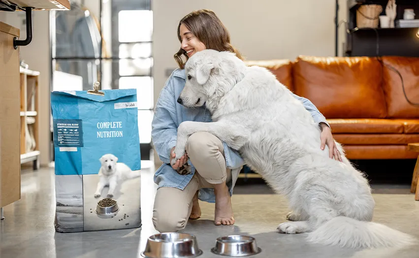 A woman kneeling holding the paw of a cute white dog in the living room. Both are looking at a bag of dog food (Photo)