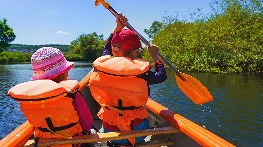 Two children who wear swim vests are rowing on a river (Photo)