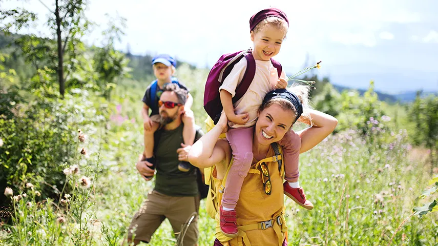 This picture shows a family of four taking a hike surrounded by nature. The two adults are carrying their children on their backs.