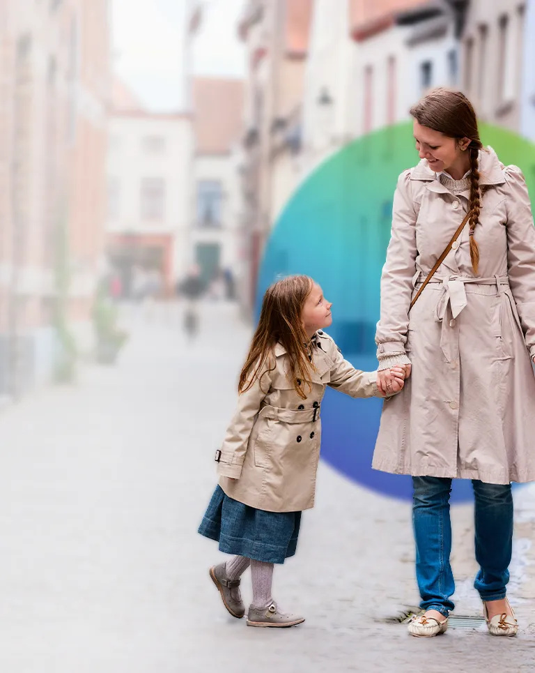 This image shows a woman and a child standing on what appears to be an old, cobblestone street. The woman is wearing a long coat and is holding the hand of the child, who also has a coat. They are both looking towards the same point, possibly engaging in conversation or observing something. In the background, there is a line of old buildings under clear sky, suggesting this photo may have been taken during daylight hours with good weather.