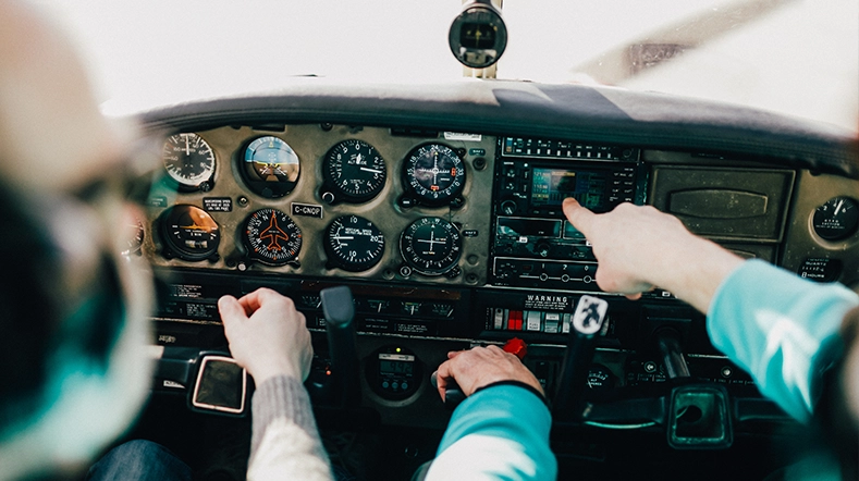 This image features an airport cockpit, showing the control panel which includes the flight instruments and flight controls. Two individuals can be seen in the image; on the left side is a cockpit with various gauges and panels, and on the right side is a person's hands pointing to these controls. The background reveals more of the aircraft, including part of the fuselage and windows for observation.