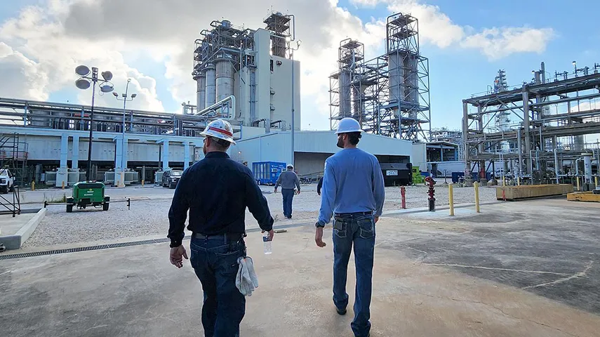 The image shows two individuals in an industrial setting, likely at a refinery or processing plant. One person appears to be engaged in conversation with the other, who is walking away.